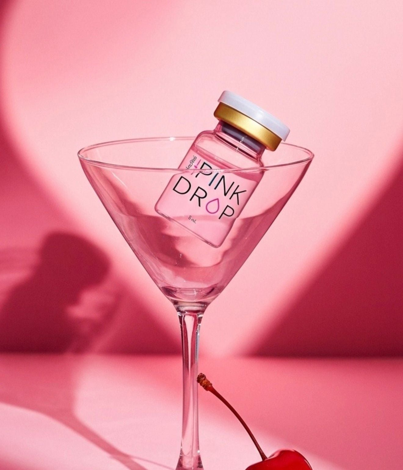 Bottle labeled 'Pink Drop' in a martini glass with a cherry on a pink background